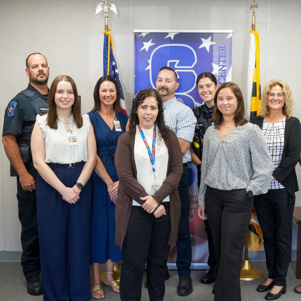 Eight people stand in front of the American flag and the Maryland state flag. Two people are law enforcement and six people are civilians.