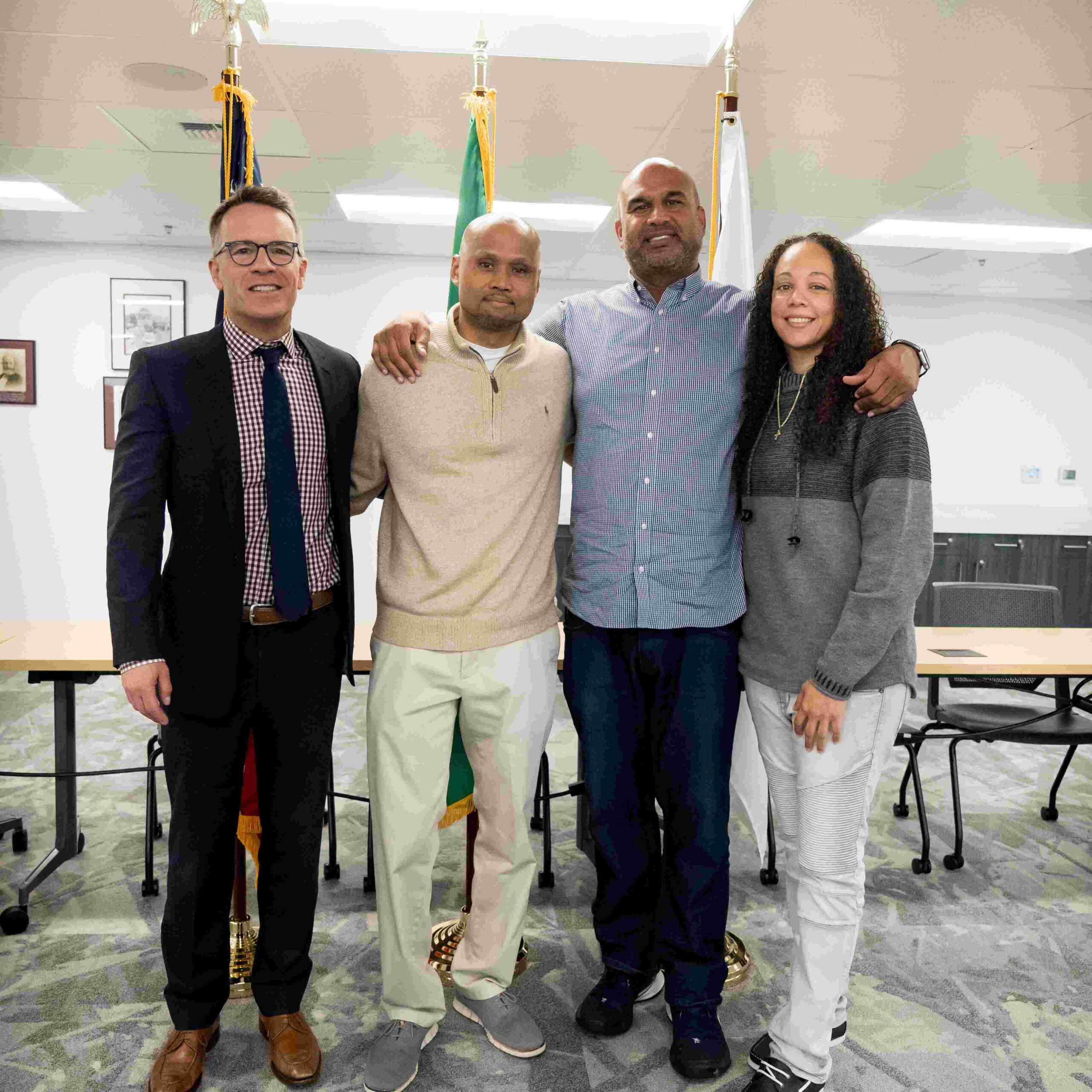 Four people standing with their arms draped around each others shoulers. They stand in front of flags in a conference room.